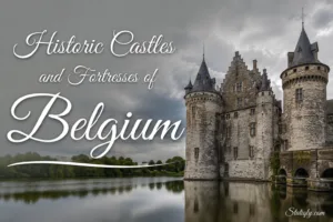 Historic castles and fortresses of Belgium sit beside a calm river with Gothic architecture and lush greenery in the bac…